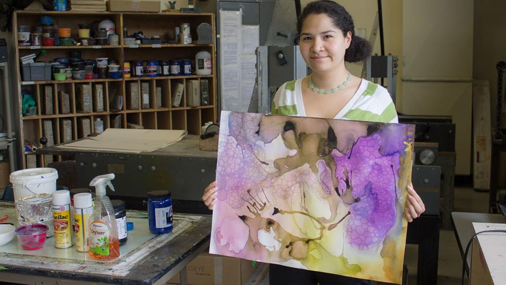 Senior Andrea Aguirre poses with an art of the piece that combines watercolor and printmaking, and will hang with the other Bachelor of Fine Arts students’ capstone portfolios in the Handwerker Gallery starting April 28. Aguirre combines her many interests and personality traits through her art. 