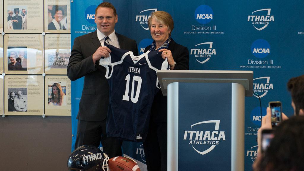 Susan Bassett  ’79, director of intercollegiate sports, hands Dan Swanstrom, new football head coach, a jersey with the No. 10 to represent him being the 10th coach in school history. 