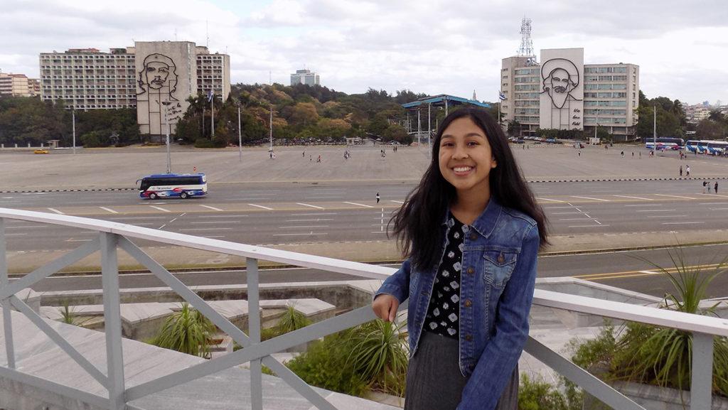 Junior Celisa Calacal poses in front of the Plaza de la Revolución, where the faces of revolutionaries Che Guevara and Camino CIenfuegos adorn government buildings.