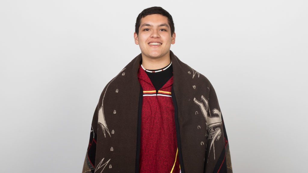 Senior Victor A. López-Carmen wears a red ribbon shirt for celebrations and is cloaked in a brown blanket from his tribe.