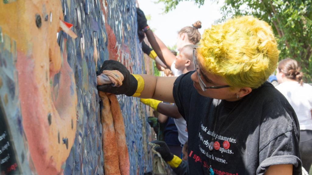 Ithaca College freshman Luis Valderrama dabs at a mosaic being created by the First Street Mosaic Project team Aug. 26. The group, composed of students participating in the Jumpstart program and community members, is crafting a mosaic mural on a wall of the downtown water and sewer Department of Public Works building.