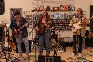 Three student band members of Lost Mary's play guitar in Angry Mom Records in front of a wall of CD's. The lead singer, Margaret, sings into the mic.