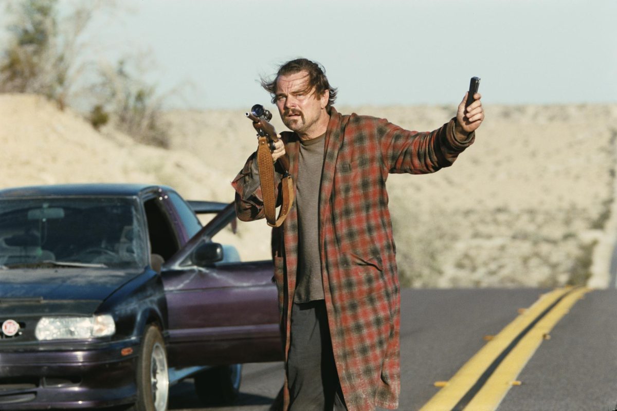 Leonardo DiCaprio stands in the street wearing a long red and black plaid jacket holding a gun by a car with an open door.