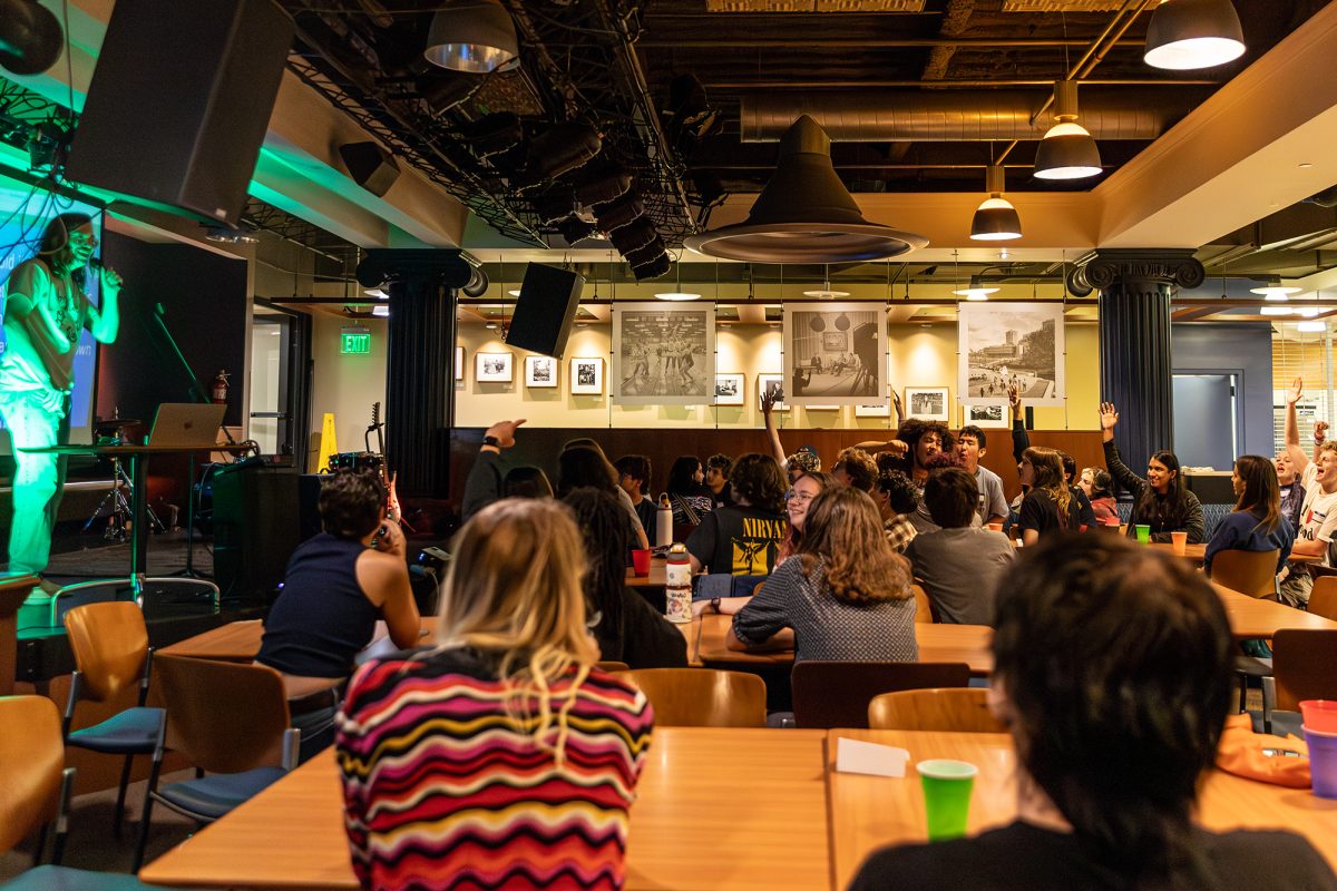 From a side angle, students sit at tables looking towards a speaker with a microphone on a small stage, some of the students are raising their hands.