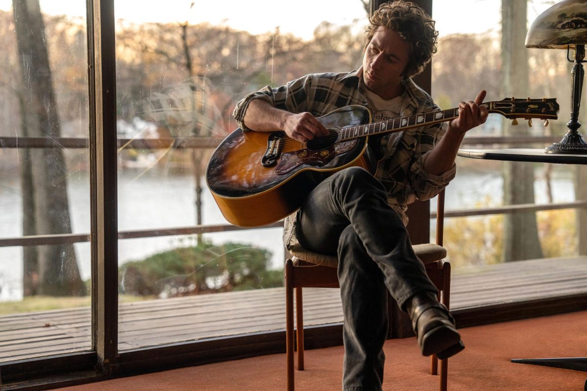 Jeremy Allen White sits with a guitar in a chair by a floor to ceiling window looking out on a lake.