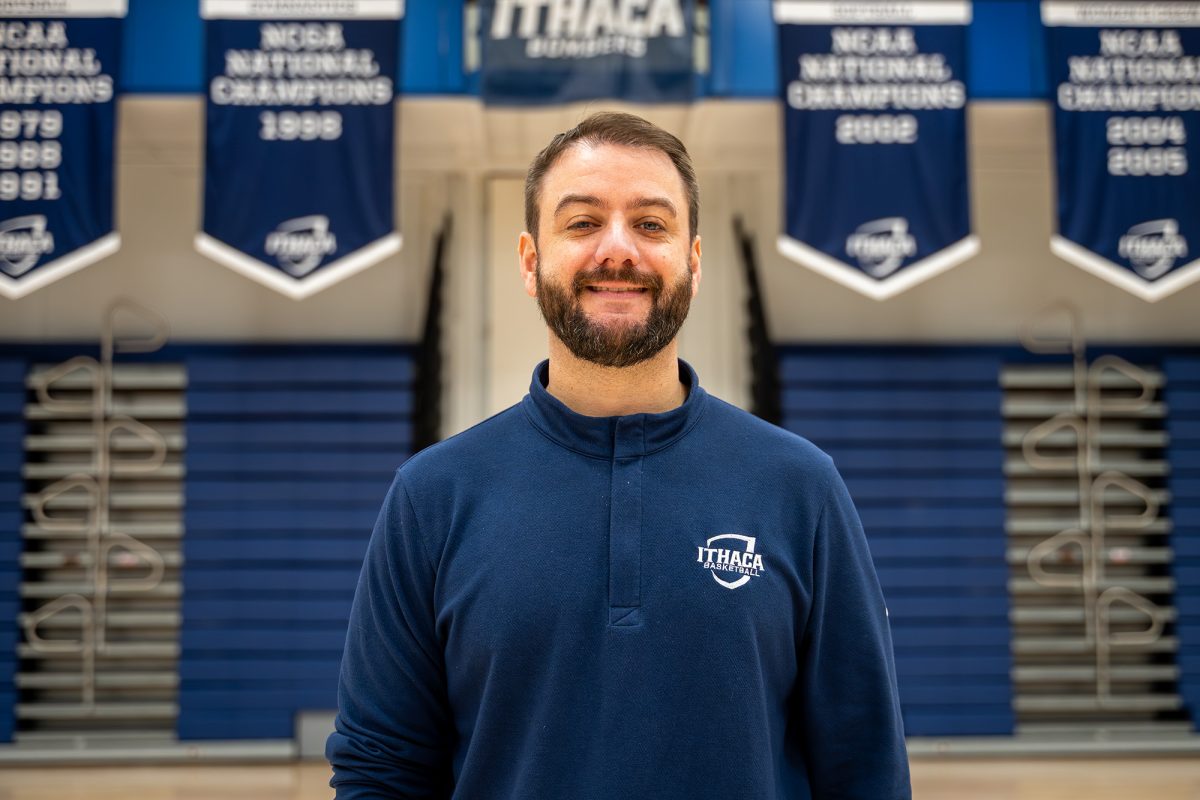 Head coach Mike McSloy stands in front of past Bomber championship banners. McSloy is entering his first season at Ithaca after coaching at Drew. 